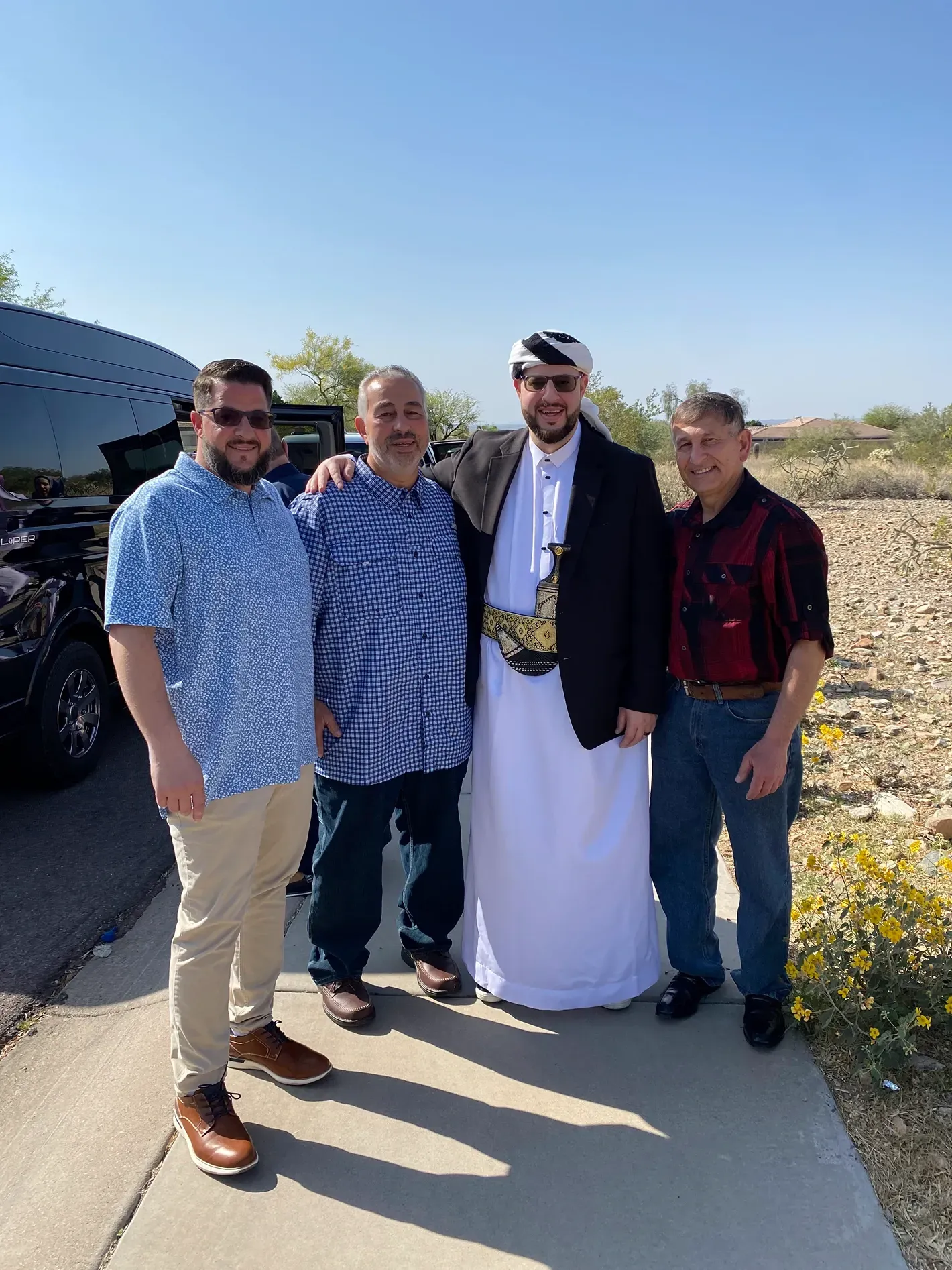 Four people standing outdoors on a sunny sidewalk, smiling beside a black vehicle.