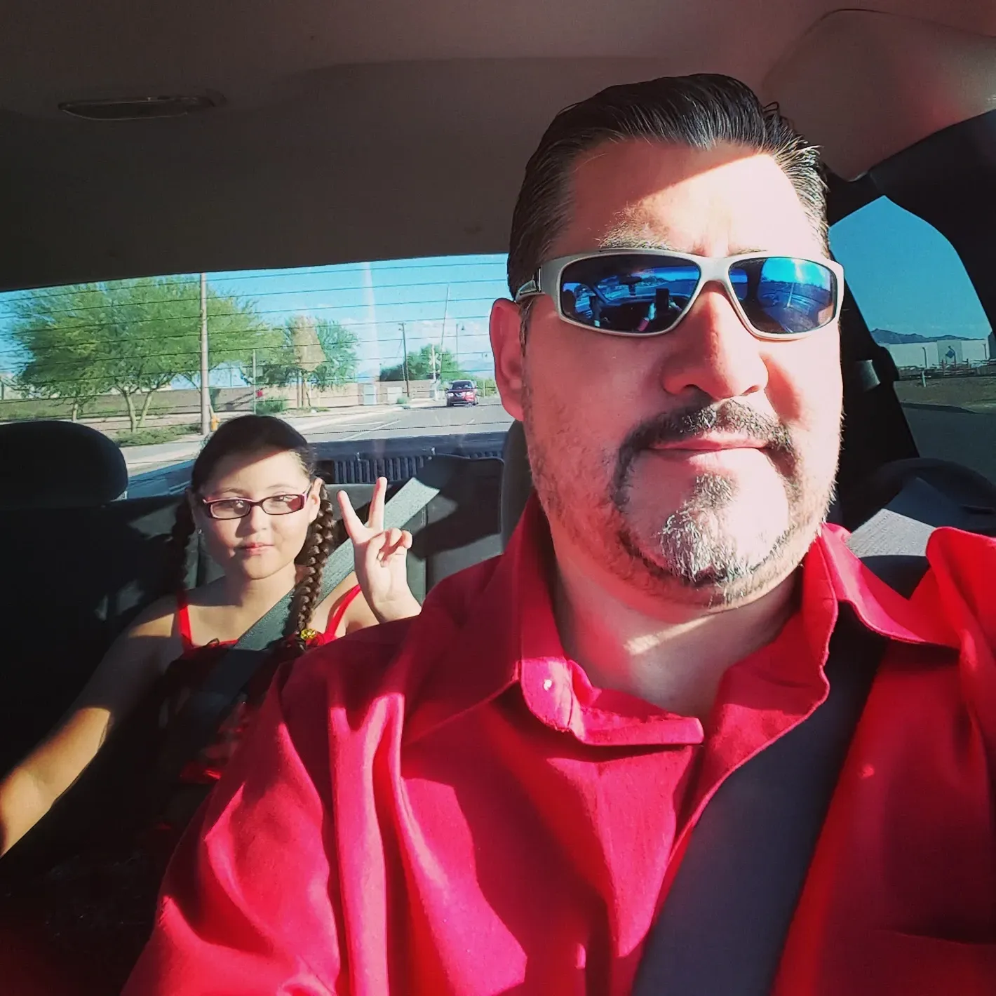 Two people in a car, front passenger wearing blue sunglasses and red shirt, backseat passenger flashing a peace sign.