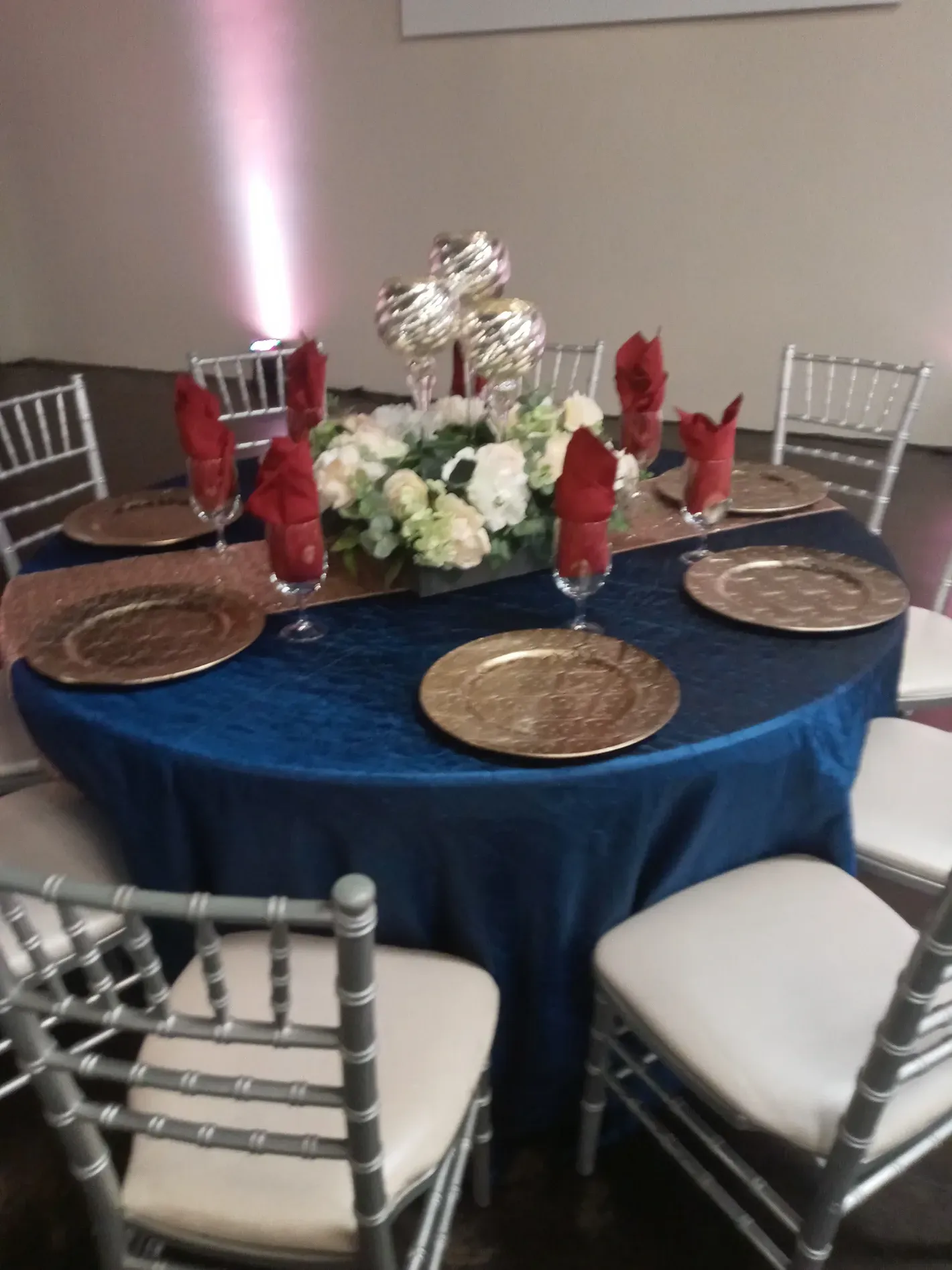 Round banquet table with navy tablecloth, silver chairs, placemats, and floral centerpiece with red napkins