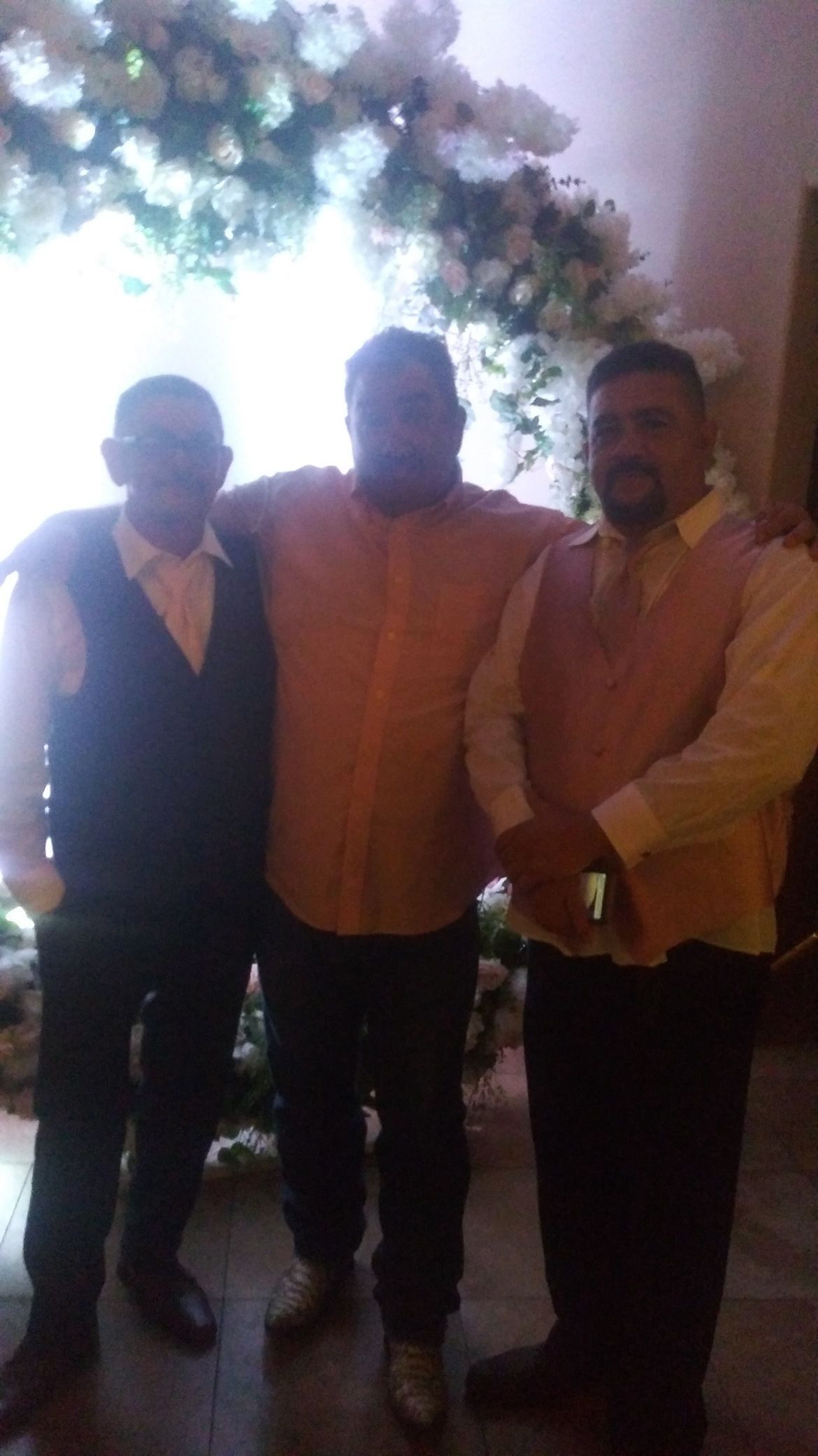 Three people posing together indoors under dim lighting, with a decorative plant overhead.