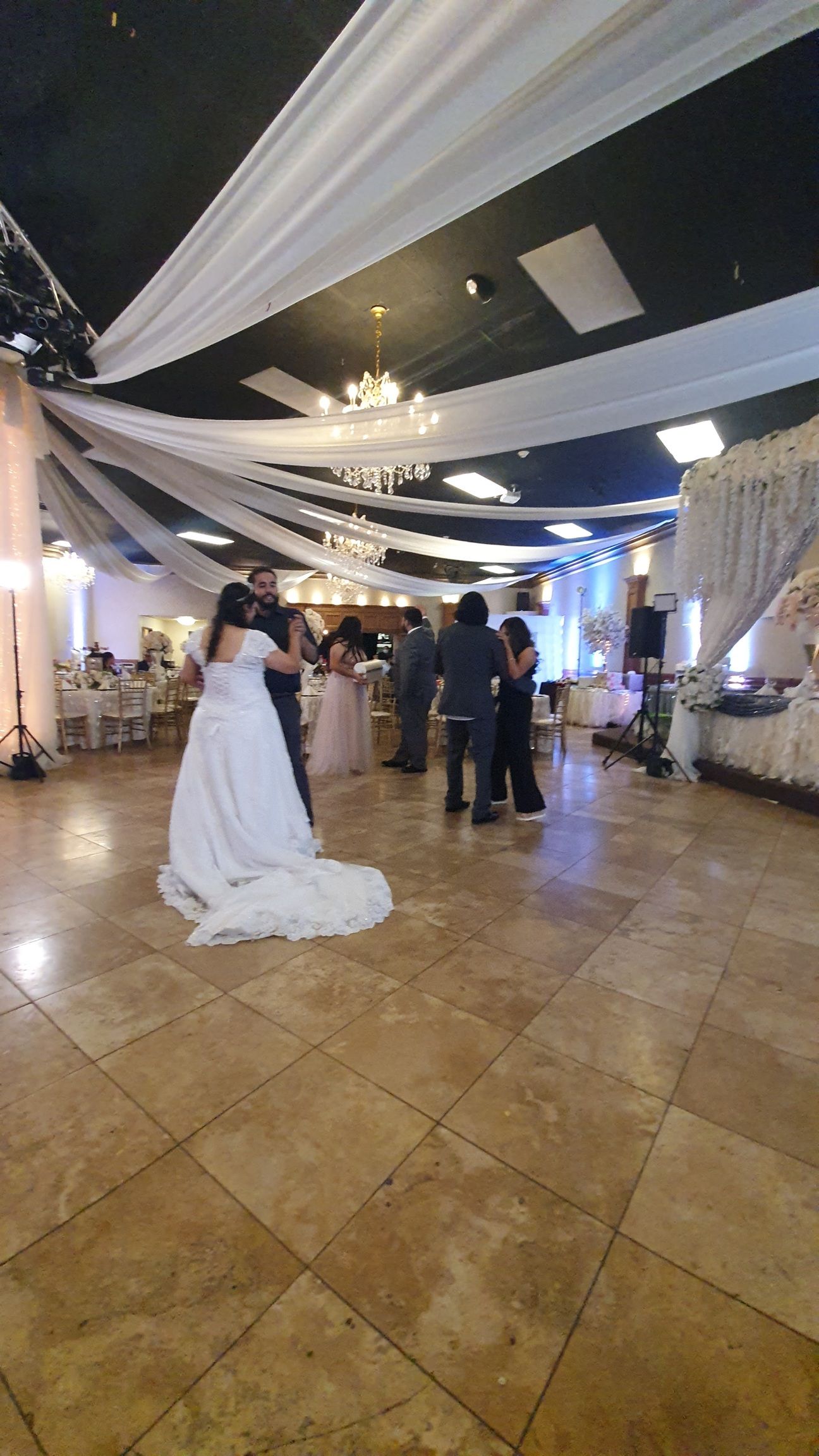 Bride in white gown on tiled dance floor at indoor wedding reception with guests and draped ceiling
