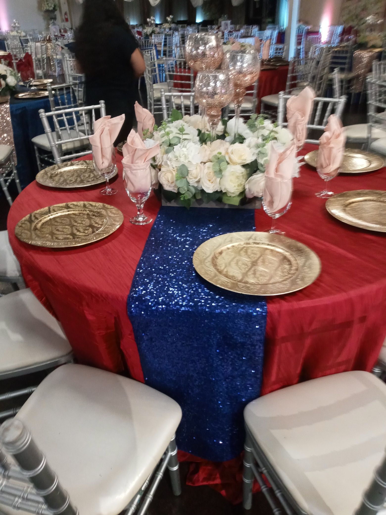 Round banquet table with red and blue tablecloth, white floral centerpiece, gold chargers, and pink napkins.