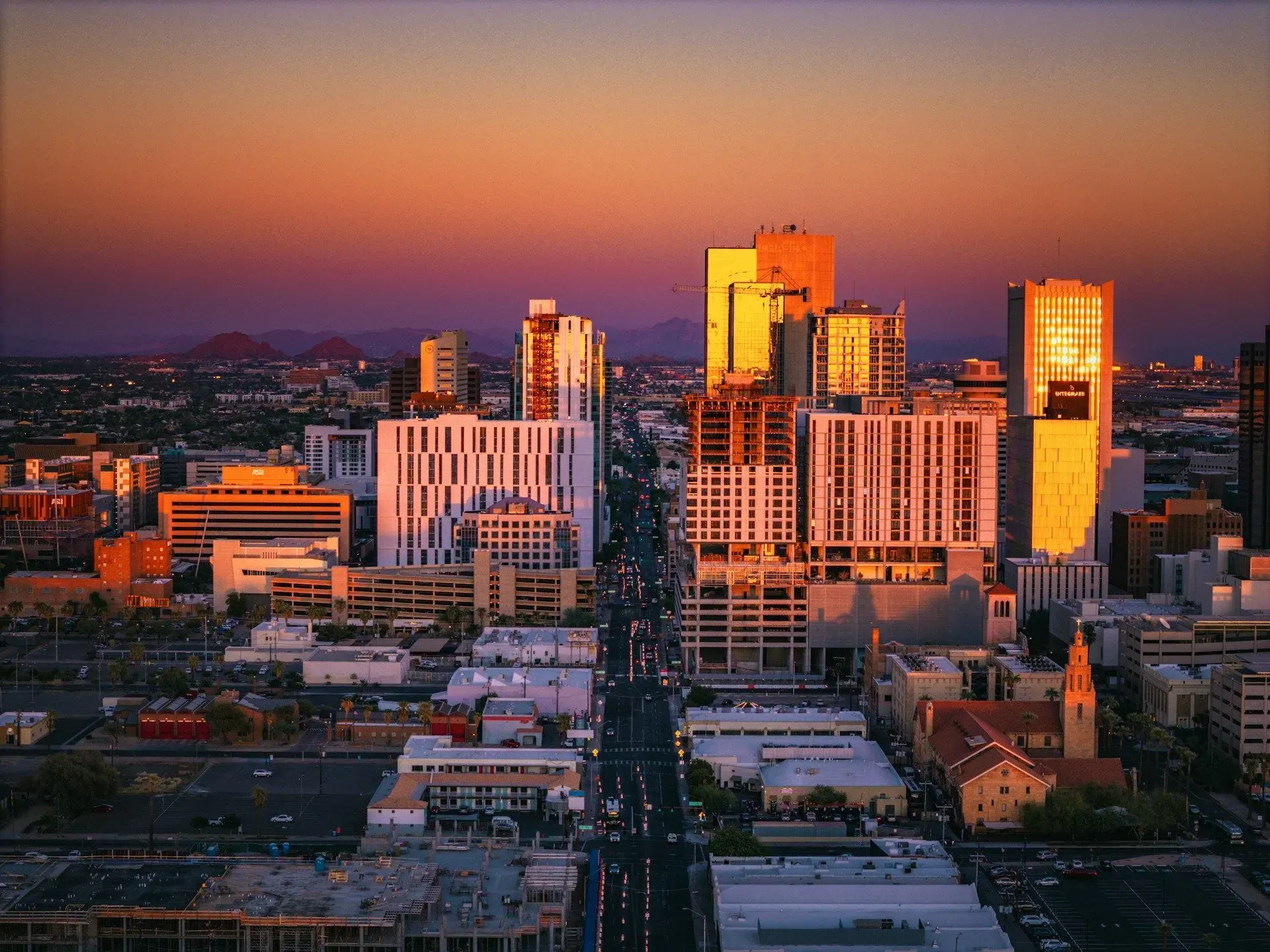 Sunset over a city skyline with tall buildings and a central road leading into downtown