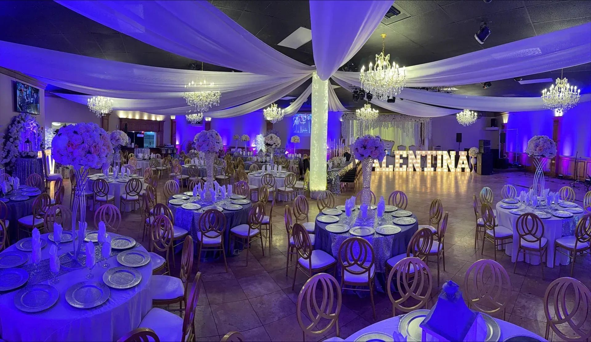 Purple-lit banquet hall with round tables, white drapes, chandeliers, and a large illuminated backdrop reading “FELIZ CUMPLEAÑOS”