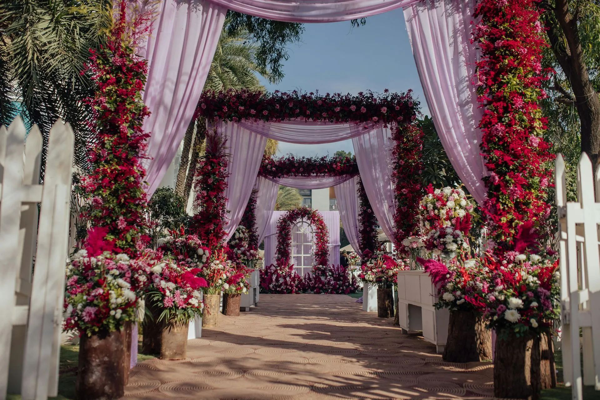 Flower-lined outdoor walkway with pink drapes and arches leading to a decorated event area
