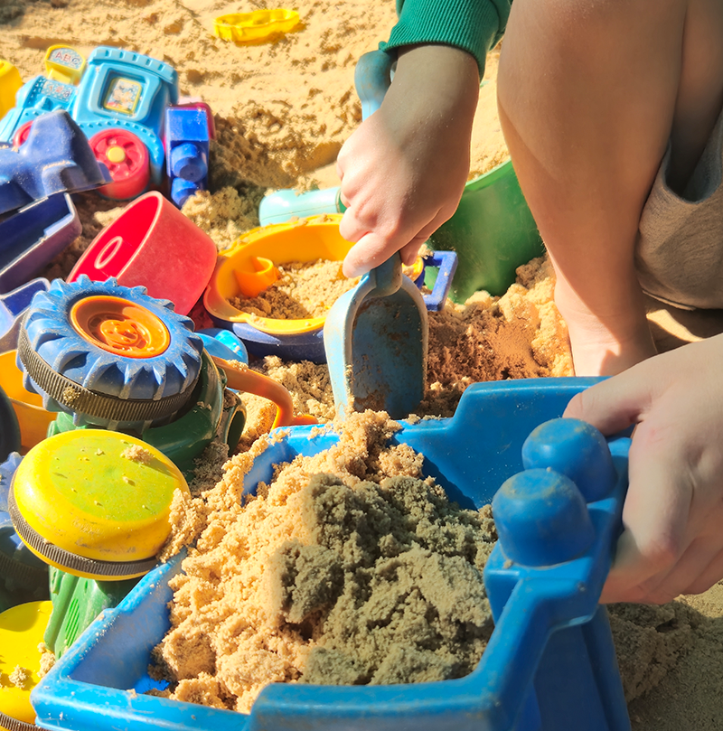 Child playing with blue dump truck and other toys in a sandbox filled with sand.