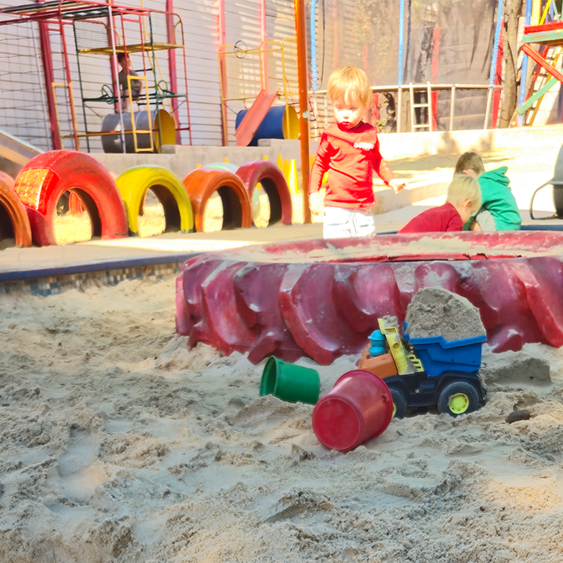 Child playing in a sandbox with toy truck and buckets. Playground equipment and other children in the background.