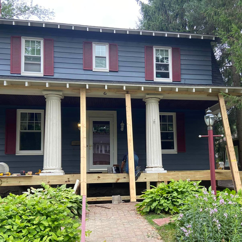 Two-story house with columns and porch under construction. Blue siding, red shutters, and wooden supports are visible.