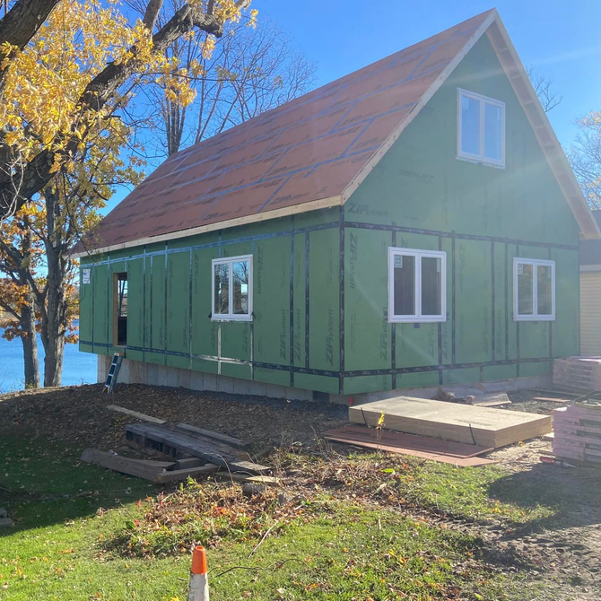Green-wrapped house under construction with white windows and a brown roof, set on a grassy waterfront, bright blue sky.