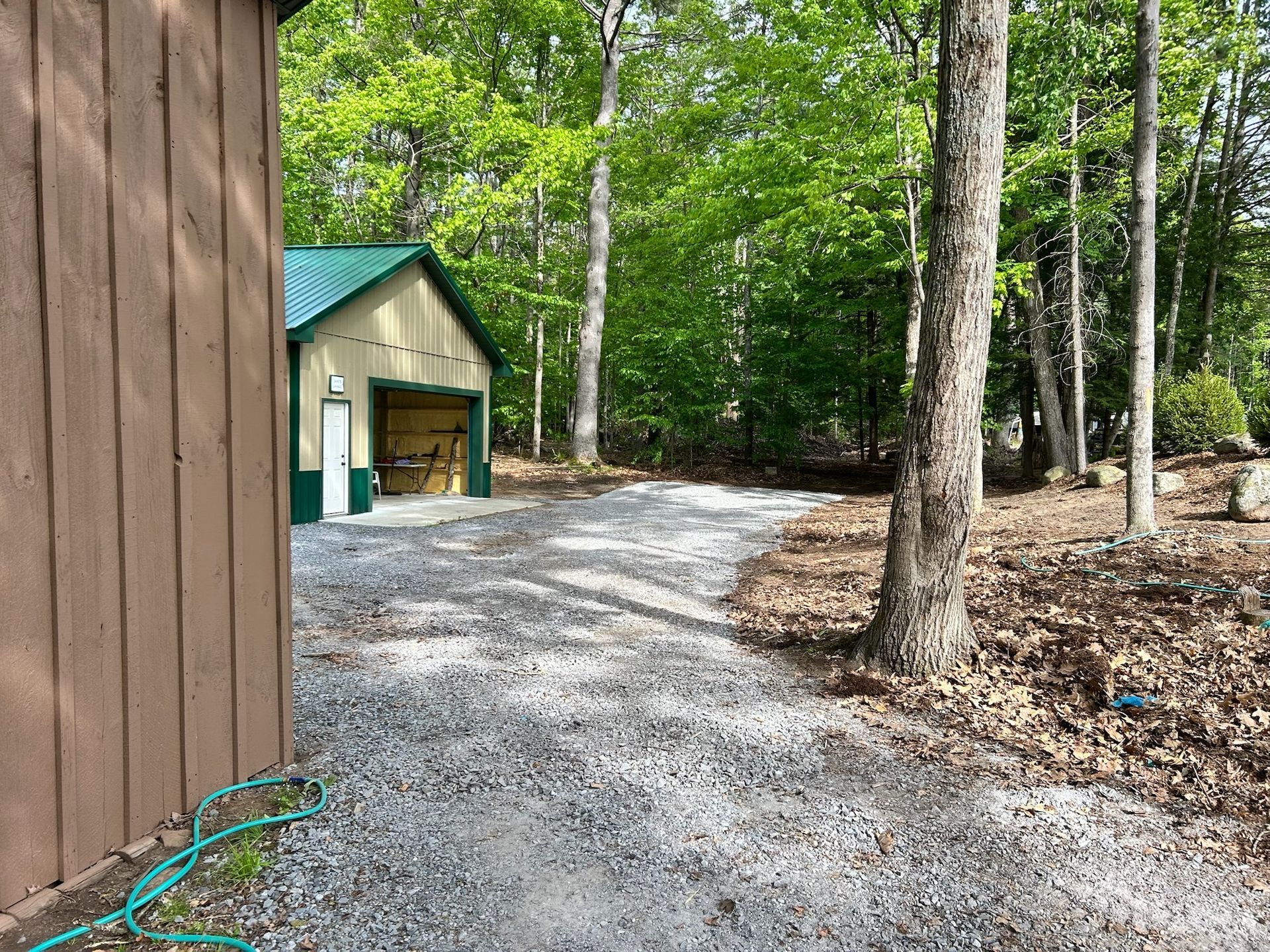 Gravel driveway beside a wooden building, leading to a small shed in a wooded area.