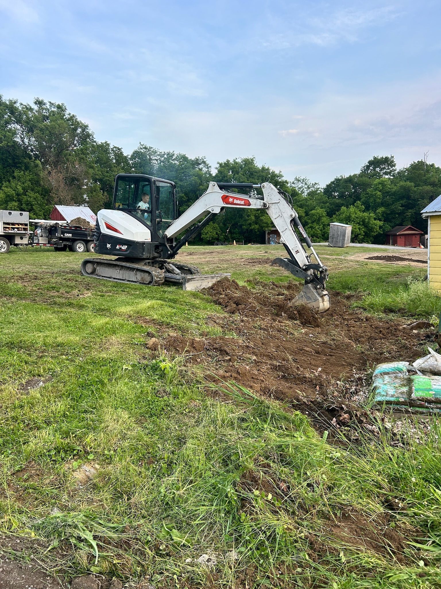 Excavator digging in a grassy field with trees and parked trucks in the background