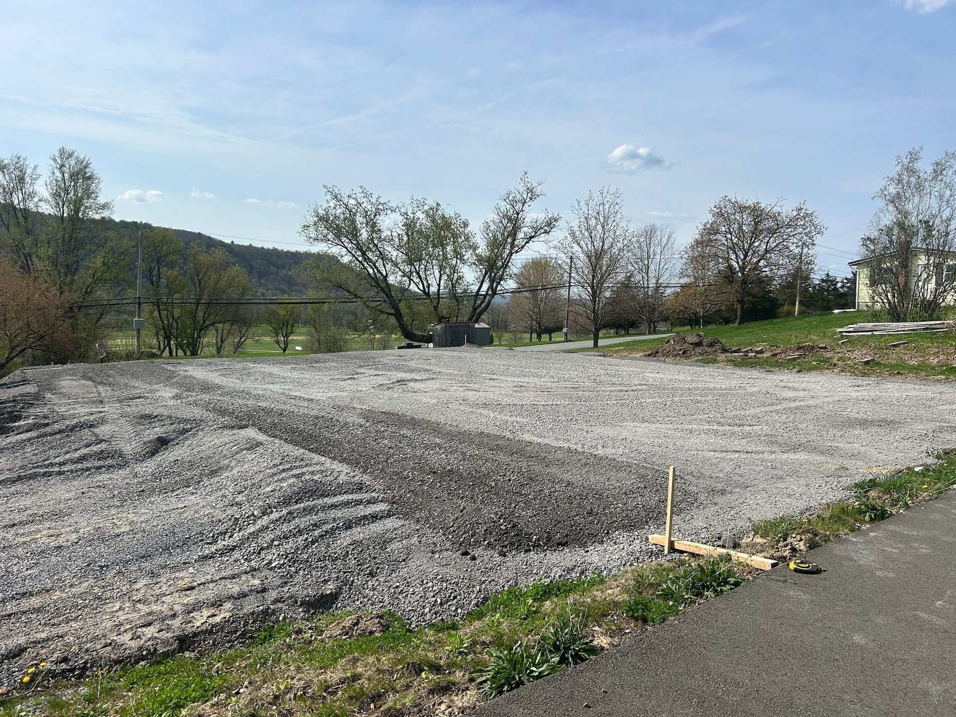 Gravel lot beside a paved road, with trees and low hills under a clear blue sky