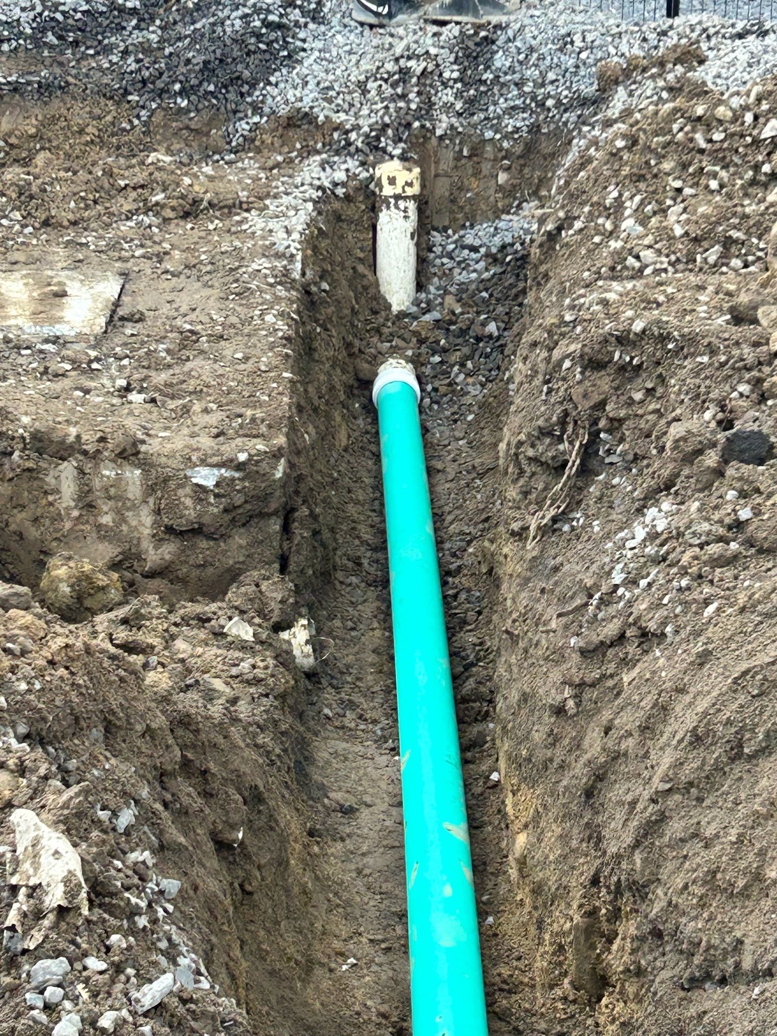 Bright green pipe in a trench beneath gravelly soil, with a white pipe section above it.