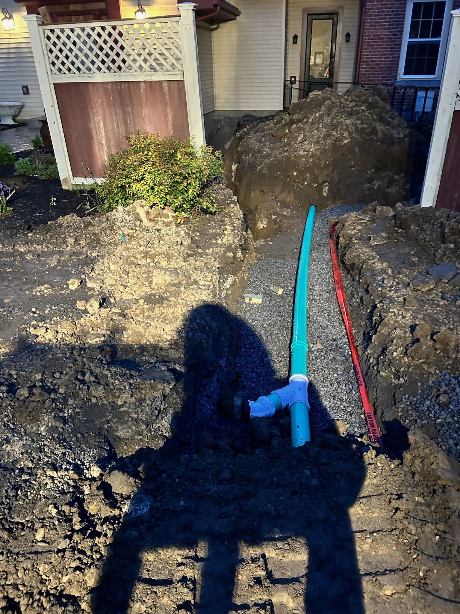 Nighttime trench beside a house, with red and blue pipes exposed in a muddy dug-out yard.