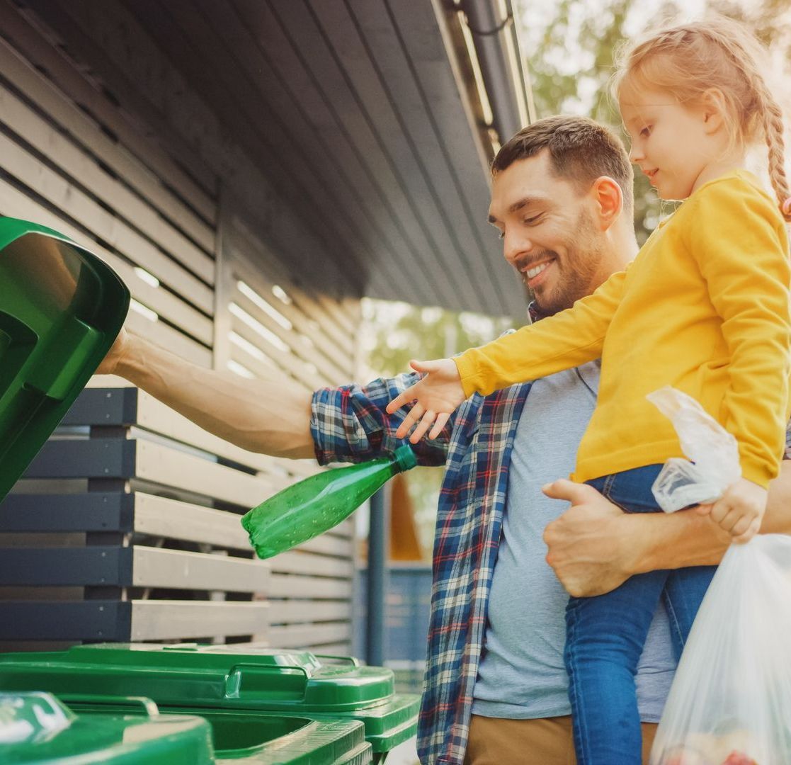 A man and a little girl are throwing bottles into a green trash can.