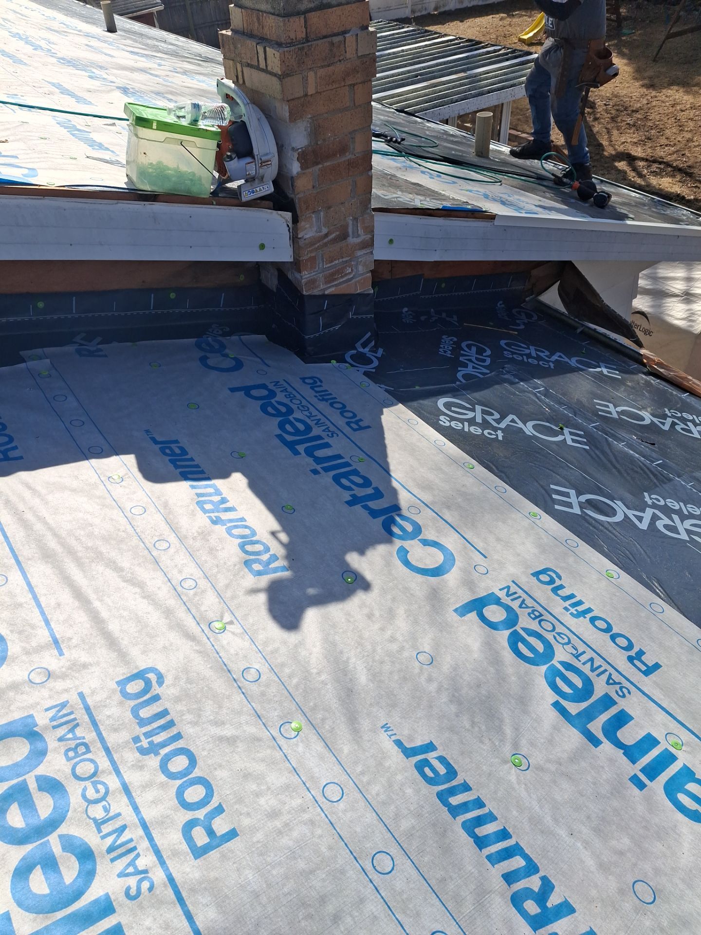 Roofer working on a roof near a brick chimney. Roofing materials and a shadow are visible.
