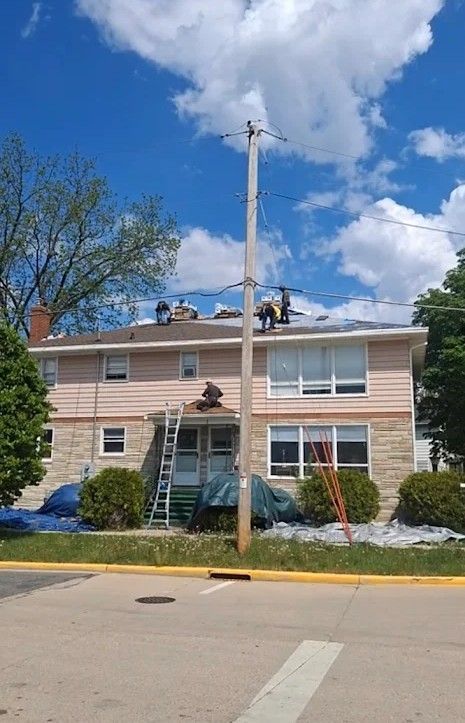Roofers working on a two-story house with a utility pole in front, blue sky, and tarp on the lawn.
