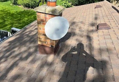 A white satellite dish is mounted on a brick chimney on a brown shingled roof, with a shadow of a person on the roof.