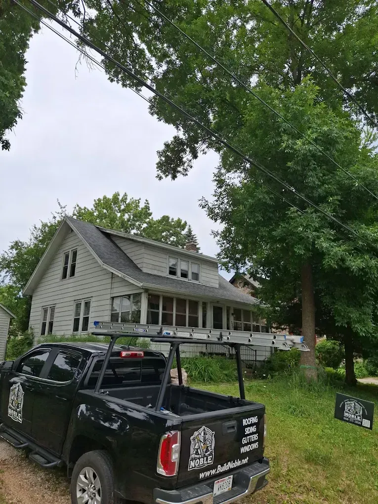 Black pickup truck parked in front of a white house with porch. Trees and power lines visible. Cloudy day.