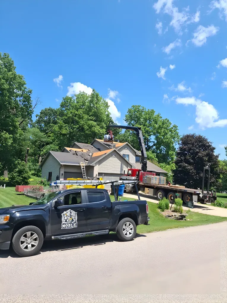 Roofers on a house with a crane; black truck in foreground, blue sky, trees.