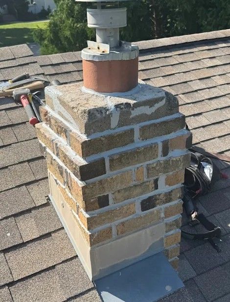 Brick chimney with metal cap on a shingled roof. Tools are visible on the roof.