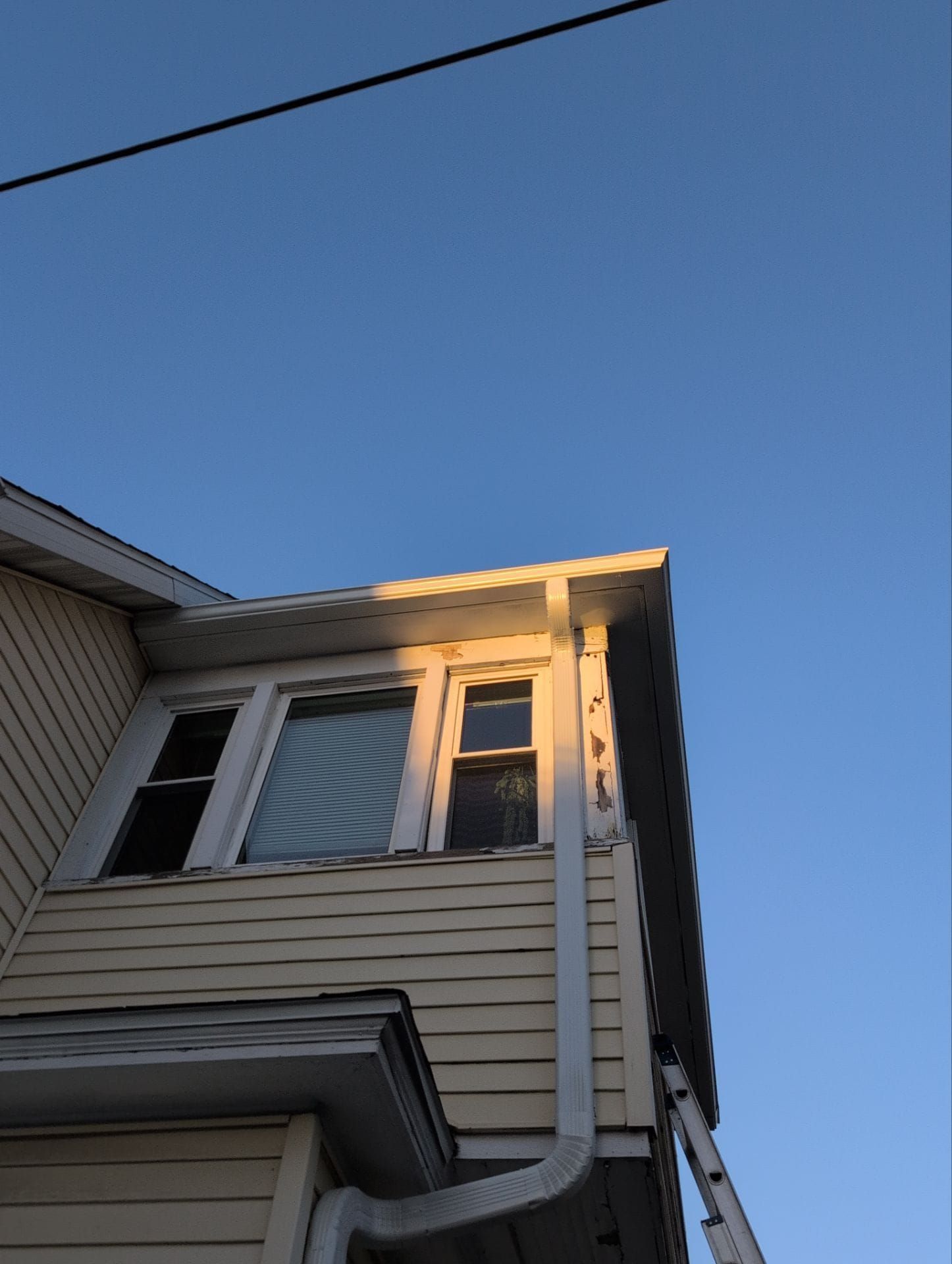 Yellow house exterior, two windows, white trim, blue sky, ladder leaning against the side.