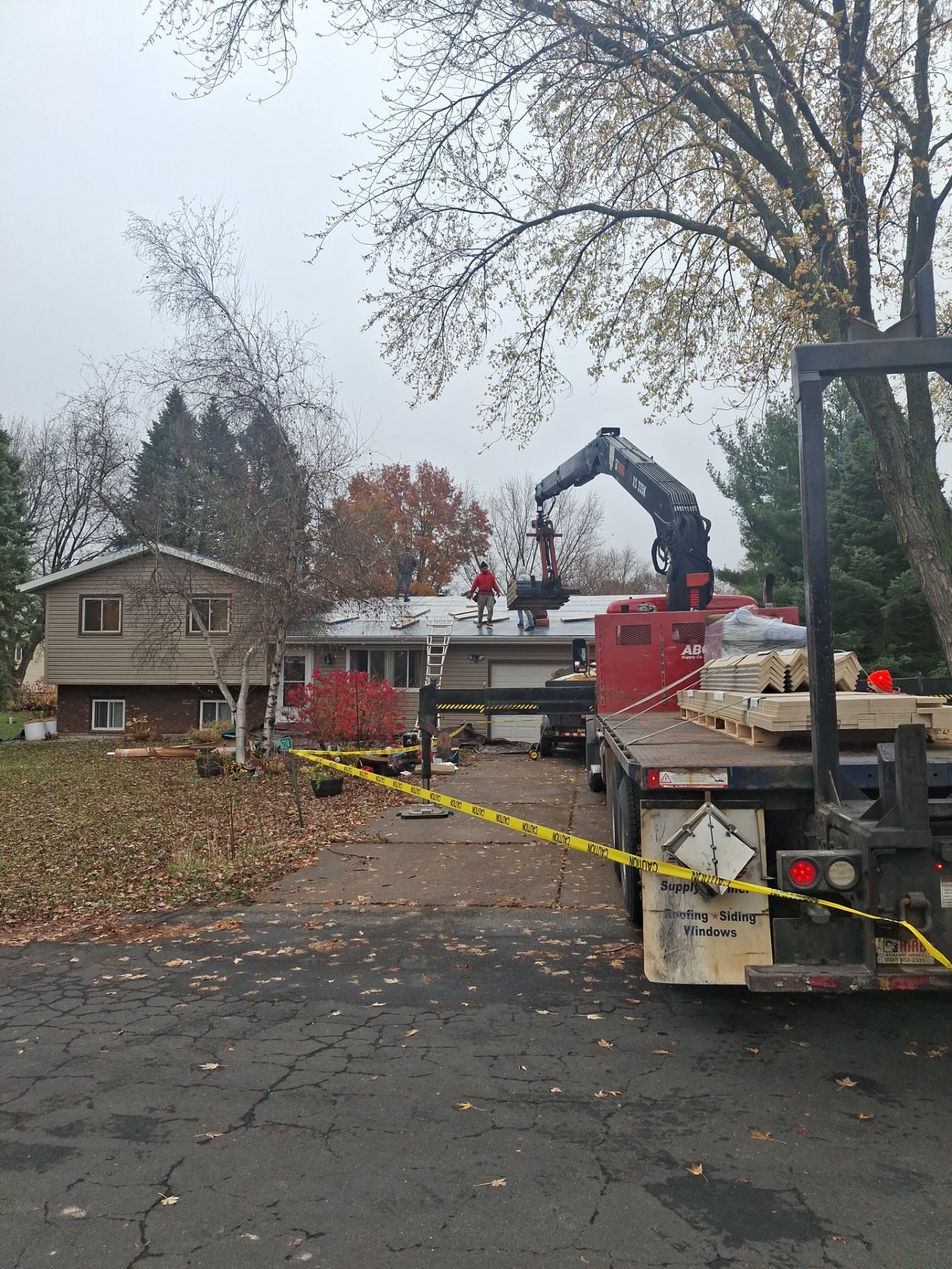 Roofers using a crane to work on a house roof; a truck with supplies is in the driveway. Cloudy day.