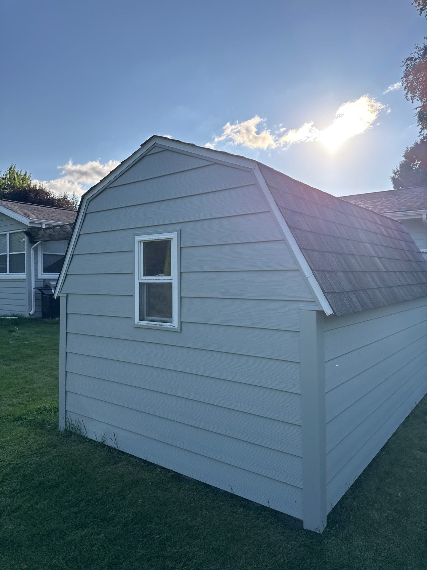 A light blue shed with a gray roof and a window sits on a grassy lawn under a sunny sky.