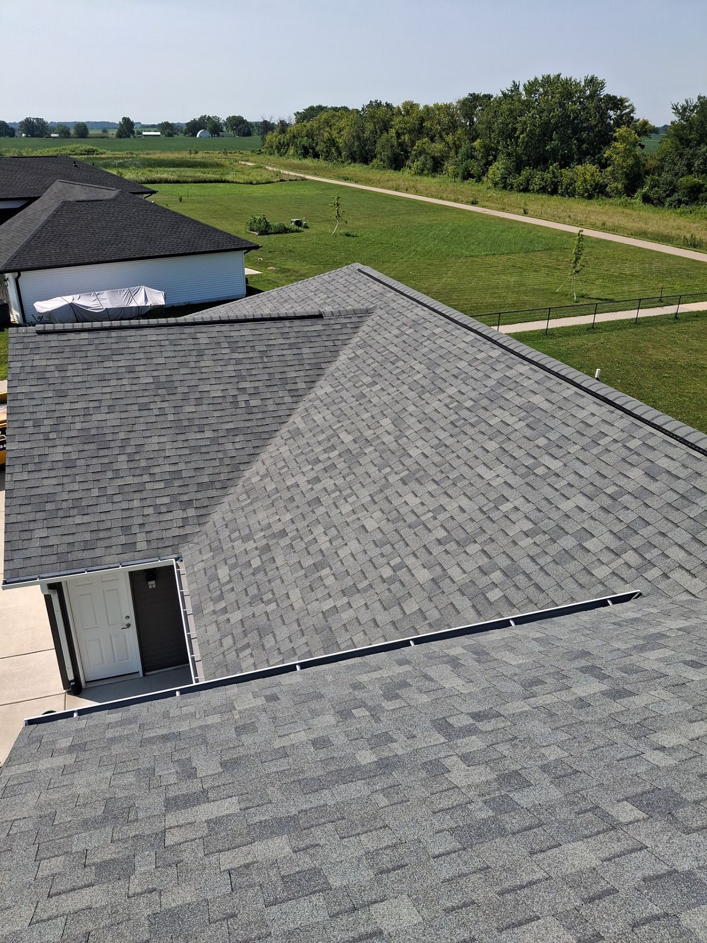 Gray shingle roof on a house, viewed from above, with fields and trees in the background under a clear sky.