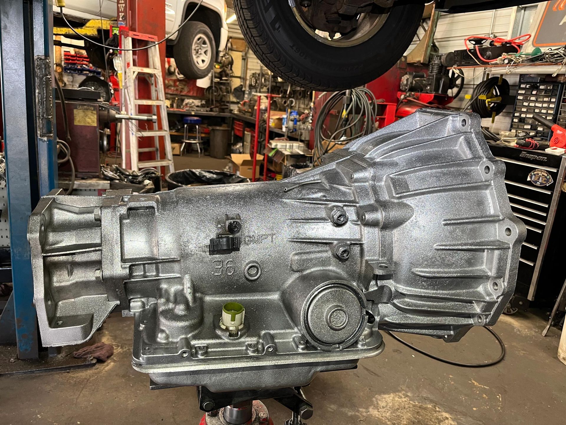 Silver-painted car transmission on a lift in a repair shop, surrounded by tools and parts.