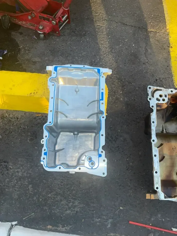 Metal engine parts on asphalt beside a yellow line, with a red tool at the top left.
