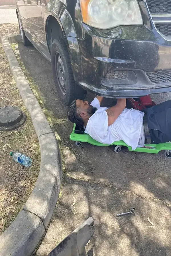 Person lying on a green creeper under a car, working on the front bumper outdoors.