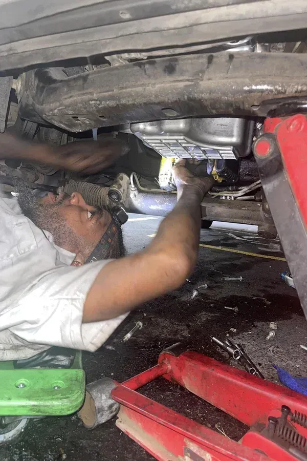 Mechanic working under a car in a garage, using a tool near the engine area.