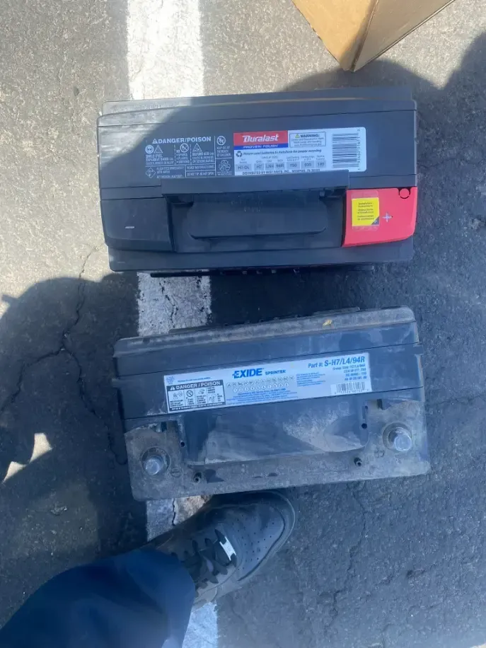 Two car batteries on pavement, one black and one dusty, with a shoe nearby and a cardboard box above.