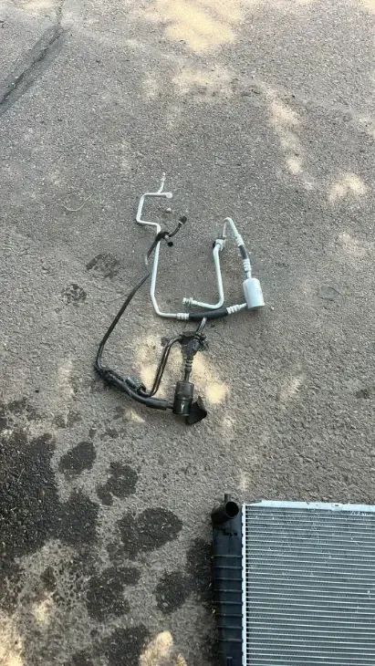 Scattered metal tools and tubing on asphalt beside a gray ribbed mat in dappled sunlight