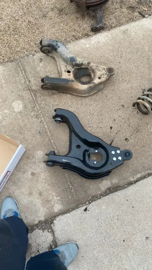 Two car suspension control arms on a concrete sidewalk, one rusty and one black.