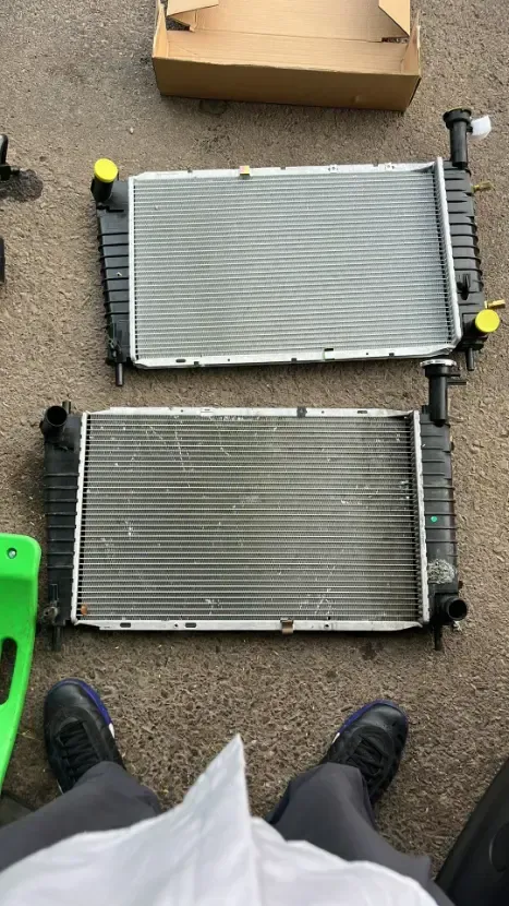 Two car radiators on a carpeted floor beside a cardboard box and a person’s shoes and legs