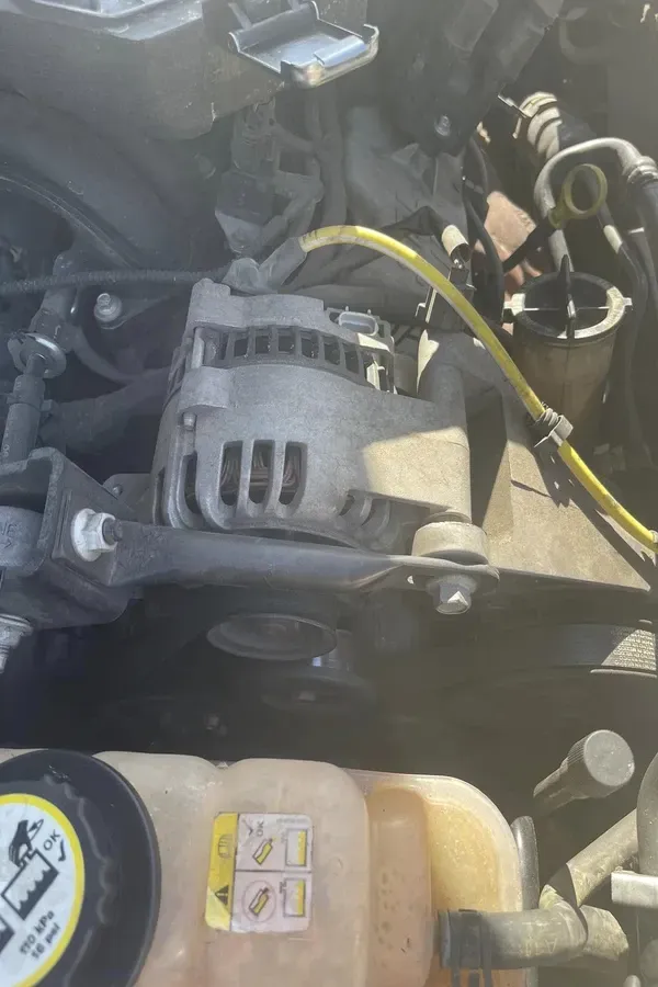 Car engine bay with alternator and yellow coolant reservoir visible, with wiring and hoses in view