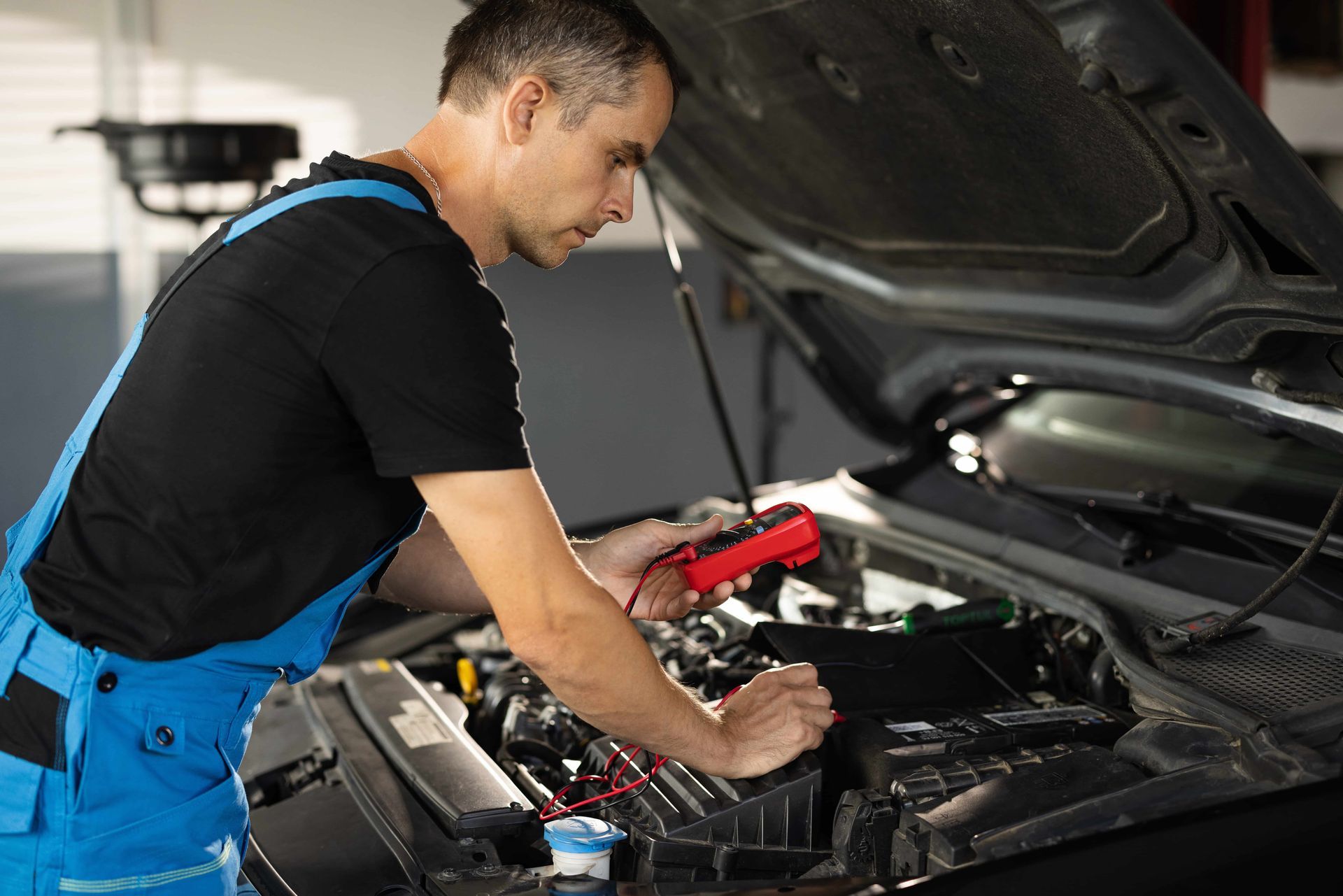 Mechanic in blue overalls working under a car hood in a garage