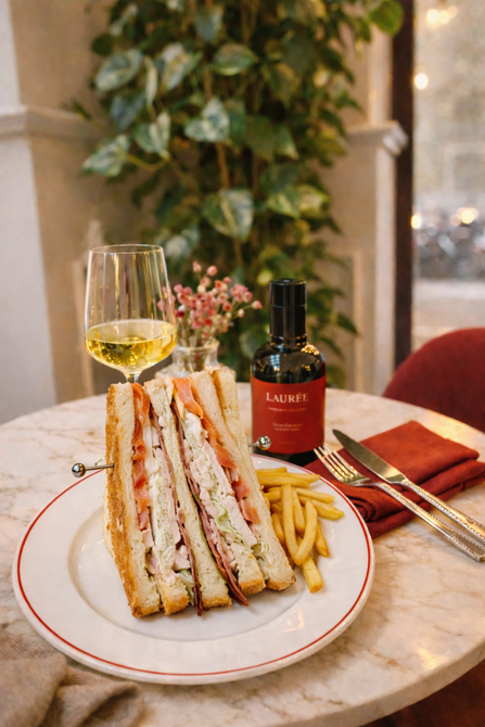 Un club sandwich e una porzione di patatine fritte su un piatto bianco, serviti con un bicchiere di vino bianco a un tavolino di marmo in un caffè.