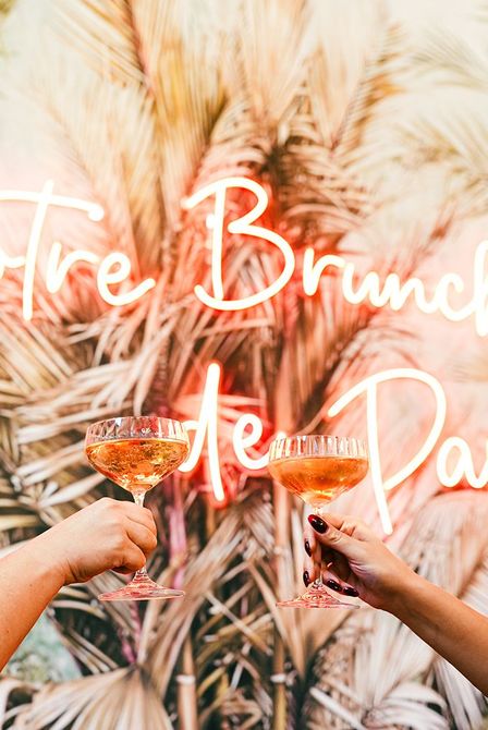 Due mani sollevano cocktail arancioni in un’ambientazione tropicale, con un’insegna al neon «Brunch» sullo sfondo
