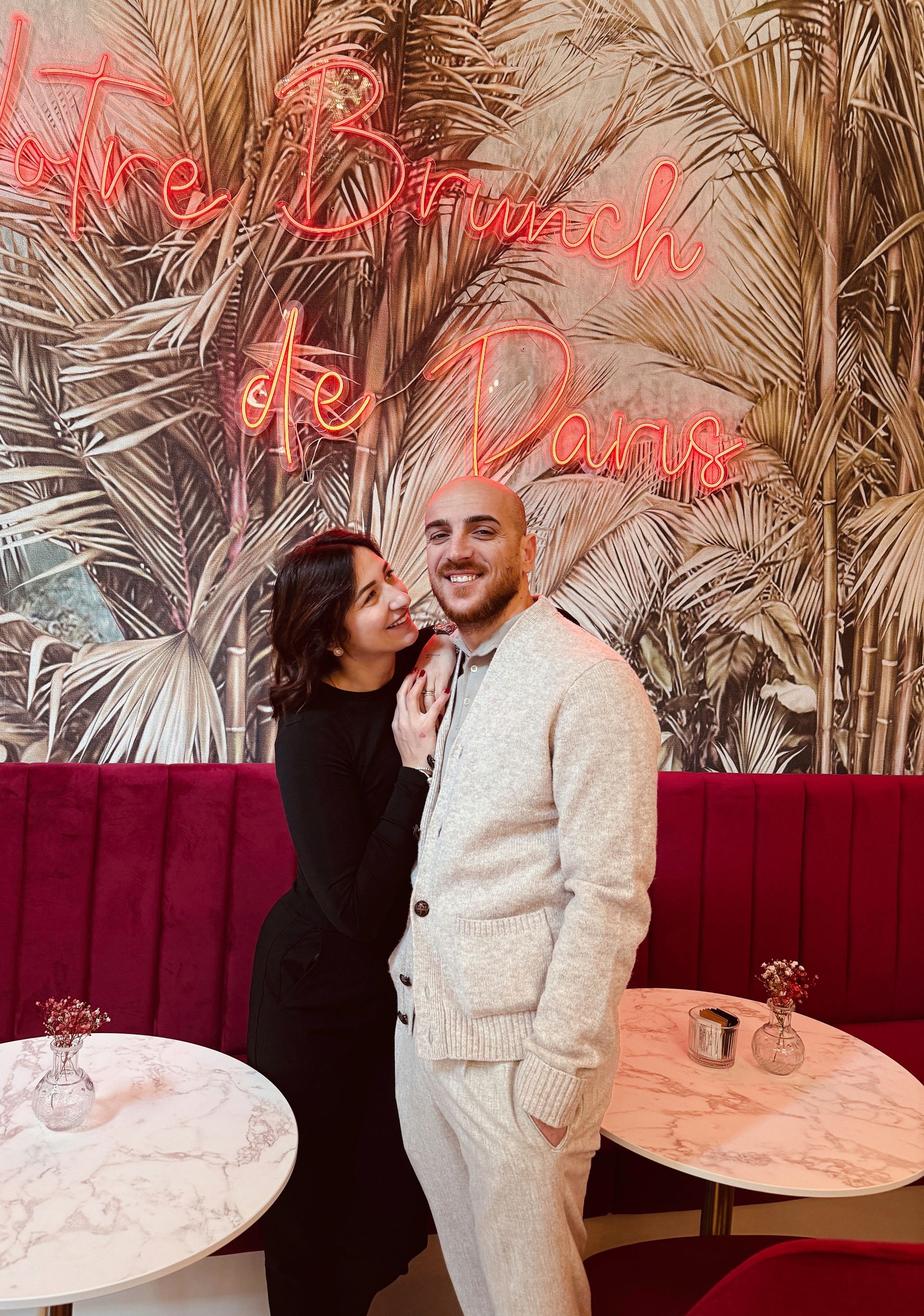 Una coppia in piedi, vicina e sorridente, davanti a un murale raffigurante foglie di palma e a divanetti rosa con scritte al neon.