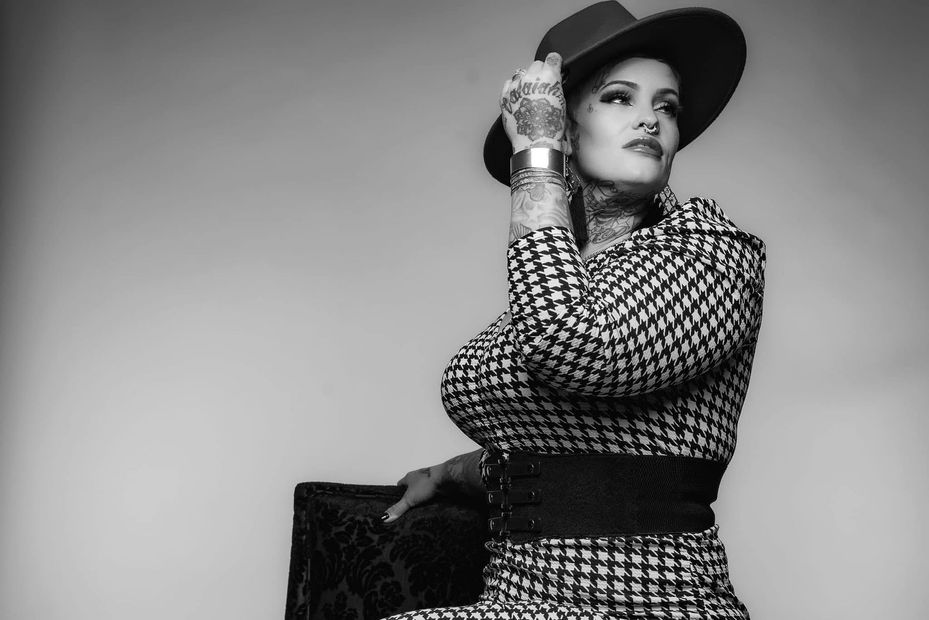 Woman in houndstooth suit and hat, hand on hat, seated. Black and white.