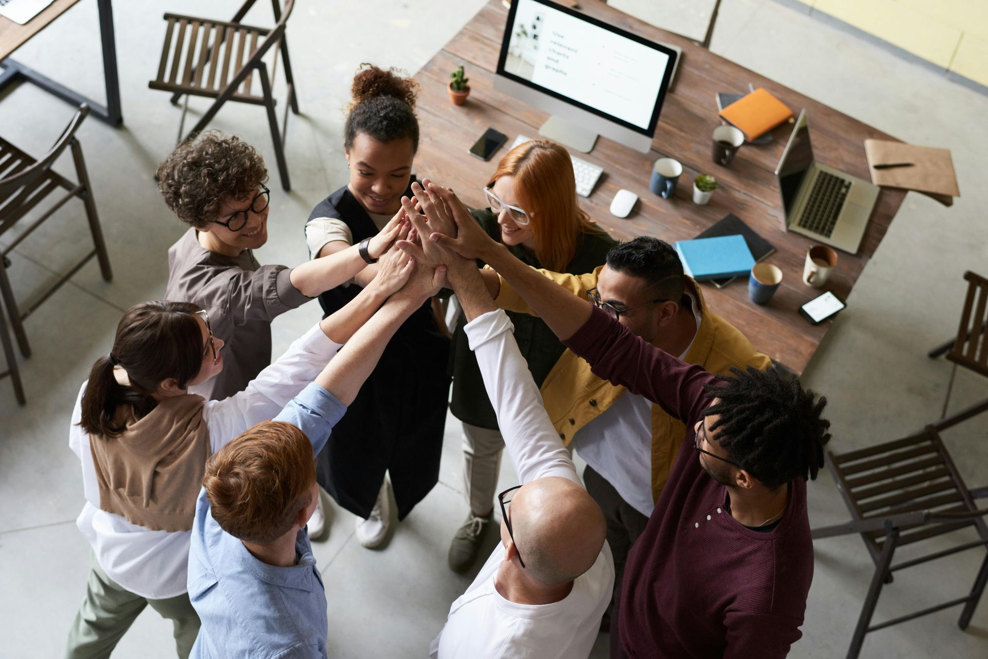 Group of people in office, hands stacked in high-five, celebrating success at a wooden table.