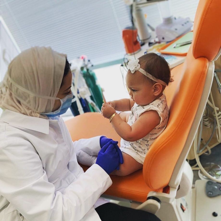Dentist in white coat and hijab examines baby in dental chair; baby holds up hand.