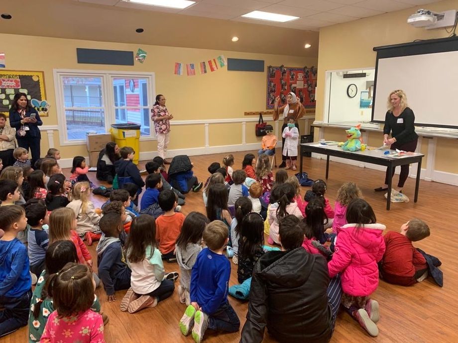 Children seated on floor, watching presenters. Classroom setting with a table displaying objects.