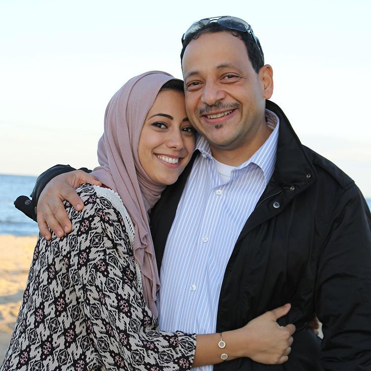 Woman in hijab and man with arms around each other smiling on a beach.