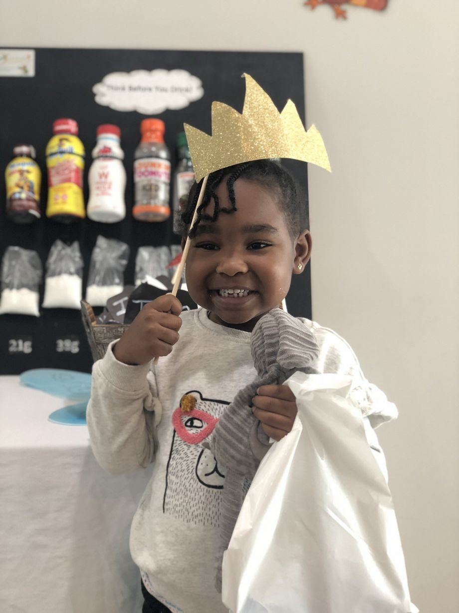 Girl wearing a crown holds a wand and bag, smiling in front of a display of ingredients and bottles.
