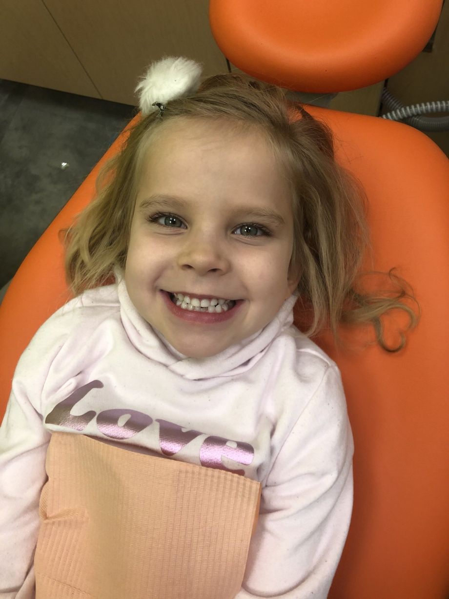 Young girl with blonde hair smiles at the dentist. She sits in an orange chair, wearing a pink shirt.