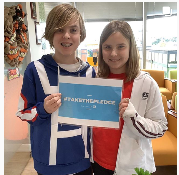 Two smiling kids holding a sign that says #TAKE THE PLEDGE. Boy: blue hoodie. Girl: red shirt, white jacket.